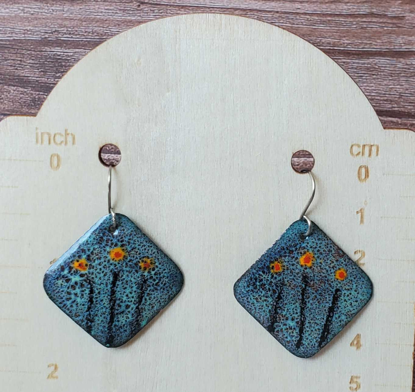Blue square copper earrings with floral design on a wooden stand with measurement scale.