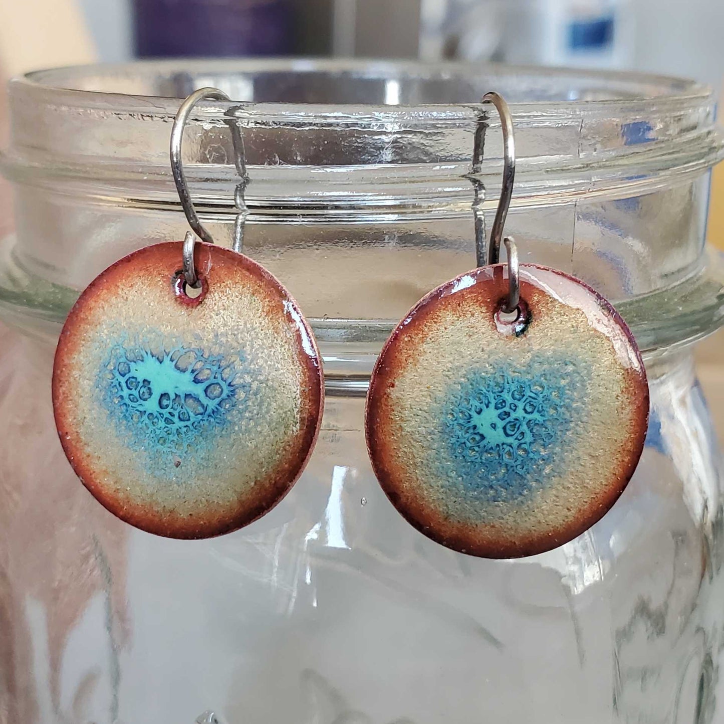 Round copper earrings with blue crackle  design hanging from a glass jar on a wooden surface.